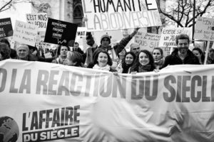 A group of people are protesting with signs in French, holding a large banner that reads "L'AFFAIRE DU SIÈCLE" during the Marche du Siècle in Paris. The signs include messages about defending biodiversity, addressing energy concerns, and responding to climate change. The photo is in black and white.