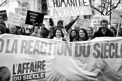 A group of people are protesting with signs in French, holding a large banner that reads "L'AFFAIRE DU SIÈCLE" during the Marche du Siècle in Paris. The signs include messages about defending biodiversity, addressing energy concerns, and responding to climate change. The photo is in black and white.