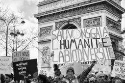 Protesters hold up signs advocating for saving humanity and biodiversity in front of the Arc de Triomphe in Paris. One prominent sign reads "Sauvons Gaia, l'humanité, la biodiversité" in French. The scene is part of the Marche du siècle, a climate change rally or demonstration in France.