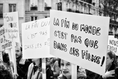 A crowd of people is holding signs during the Marche du siècle protest in Paris. The signs are in French, with messages like "LA FIN DU MONDE C'EST PAS QUE DANS LES FILMS," which translates to "The end of the world is not just in the movies." The scene is captured in black and white.