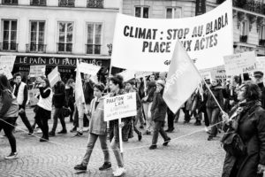 A black-and-white photo showing a group of people marching in a protest in Paris. They are holding various signs, one of which prominently reads, "CLIMAT: STOP AU BLABLA, PLACE AUX ACTES." The scene is set on a cobblestone street with buildings in the background, reminiscent of the Marche du siècle.