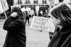A black-and-white photo captures a street protest in Paris. A person holds a sign reading "Liberté, Égalité, Potager" in French (Liberty, Equality, Garden). Other demonstrators hold various signs. The background features a shop labeled "Le Pain du Bourg" and cobblestone pavement.
