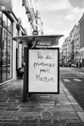 Black and white image of a city street in Paris with a glass bus stop shelter in the foreground. The shelter has a handwritten message on it that reads, "Pas de printemps pour Macron." Buildings and pedestrians are visible in the background, capturing a moment from the Marche du siècle.