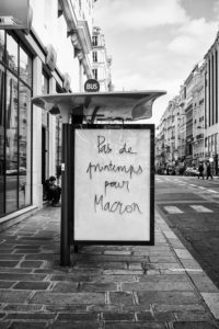 Black and white image of a city street in Paris with a glass bus stop shelter in the foreground. The shelter has a handwritten message on it that reads, "Pas de printemps pour Macron." Buildings and pedestrians are visible in the background, capturing a moment from the Marche du siècle.