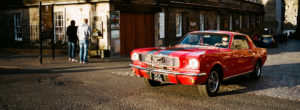 A classic Ford voiture rouge with white racing stripes is parked on a cobblestone street in front of an old stone building. In the background, two people are standing and facing a notice board on the wall. The scene is bathed in warm sunlight.