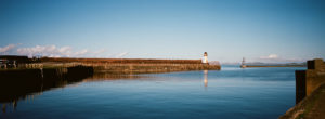 Panoramic view of a calm harbor with clear blue water. Le phare is situated on a long pier extending into the water. Another distant structure stands further into the sea. The sky is clear with a few scattered clouds. A railing runs along the left side.