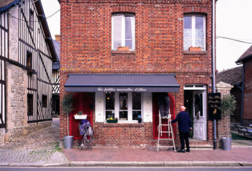 A street view of a brick building with a store named "Les petites merveilles d'Alice" on the ground floor. A person stands on a step ladder adjusting items on the store’s display, which features clothing and accessories. The street is lined with half-timbered houses.