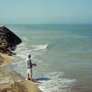 A person wearing a hoodie and shorts is standing on a sandy beach, fishing with a rod. Gentle waves lap at their feet, reminiscent of Le pêcheur de Villerville, and a rocky breakwater extends into the calm sea under a clear blue sky.