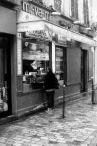 Black-and-white photo of a person standing in front of a falafel casse-croute vendor named "mi-va-mi." The shop's sign advertises "The Best Of The Street" falafel. The person appears to be ordering as they face the vendor, and the setting is on a cobblestone street.