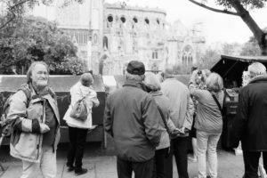 A foule of people, some taking photos, are gathered in front of a Gothic-style cathedral. Trees partially frame the scene. One man in the foreground looks at the camera, while others focus on the architectural details of the cathedral in the background.