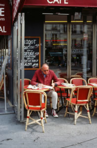 A man in a red jacket and gray pants sits alone at a small outdoor café table, reading a menu. The café, adorned with red and white wicker chairs and a blackboard menu beside the entrance, radiates the charm of "La vie en rouge." The setting appears to be on a bustling city street.