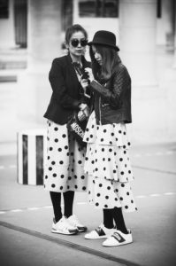 Two women dressed in matching robes à pois skirts, black jackets, and sneakers stand together on a city street. One woman wears a wide-brimmed hat and leather jacket, while the other sports large sunglasses and holds a bag labeled "Moschino." They are looking at a phone.