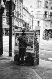 A person dressed in a jacket and cap, resembling *Le colleur d'affiche*, is posting or examining a flyer on a weathered poster board on a city sidewalk. In the background, people walk near shops and buildings at a street intersection. The scene is in black and white.