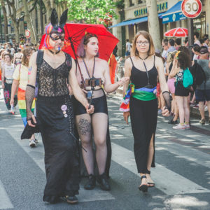 Three people are walking in a 2019 Gay Pride parade. One wears a black lace dress, fishnet stockings, and a bunny-eared mask. Another has a red umbrella, a bare chest, and tattoos. The third is in a black dress with rainbow accessories. Other parade attendees are in the background.