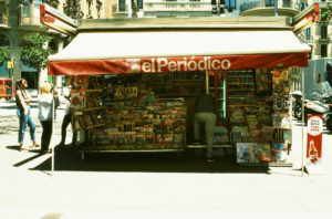 A newsstand on a sunny street in Barcelone, with a red awning displaying the sign "El Periódico." Various magazines, newspapers, and other publications are on display. Three people are near the kiosque: one browsing, one engaged in conversation, and one walking away.