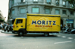 A yellow delivery truck emblazoned with the "Moritz Barcelona" logo is parked on a city street. The backdrop showcases a classic stone building with rounded corners and multiple windows, while various motorcycles are parked along the sidewalk.