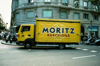 A yellow delivery truck emblazoned with the "Moritz Barcelona" logo is parked on a city street. The backdrop showcases a classic stone building with rounded corners and multiple windows, while various motorcycles are parked along the sidewalk.