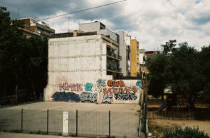 An urban area in Barcelone's Sants district showcases a blank concrete building prominently displaying vivid graffiti art. The graffiti includes colorful tags like "NITER" and "EGOR." The surrounding area features trees, residential buildings, and a fenced playground. The sky is partly cloudy.
