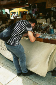 A person wearing a striped shirt and jeans is bending over a table at an outdoor market. The table, covered with a beige cloth, displays various items that look like nouvelle merveilles, including books and trinkets. The person has a black bag slung over their shoulder.