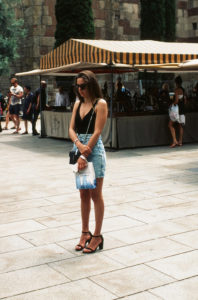 Elisa Roger, a woman with long hair, wearing sunglasses, a black top, a denim skirt, and black sandals, stands in a plaza holding a shopping bag. Behind her, there is a market stall with a striped canopy and several people browsing the goods.