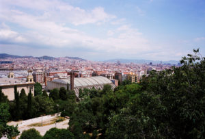 An expansive view of a cityscape under a blue sky with few clouds, featuring a mix of modern and historic buildings, lush greenery in the foreground, and distant hills in the background. A prominent domed structure stands out in the scene, reminiscent of Barcelone's majestic Parc de Montjuïc.
