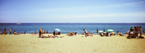 A sunny beach scene in the Barceloneta Neighborhood with numerous people enjoying the day. Some are lying on towels while others are standing or sitting under colorful umbrellas. The clear blue sea stretches out to the horizon with sailboats visible in the distance. The sky is clear.