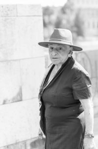 Black and white photo of an elderly person wearing a wide-brimmed hat, a short-sleeved dress, and earrings. They stand in front of a stone wall, looking directly at the camera with a neutral expression. The image captures Sabine Weinstein's quiet dignity against the rustic backdrop.