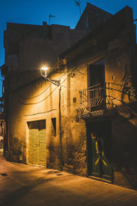 A narrow, dimly-lit street at dusk in Tarragone reveals an old building with yellow metal shutters and a balcony. A single lamp post casts a warm glow, highlighting the worn texture of the walls. Shadows create a mysterious ambiance in this nuit scene.