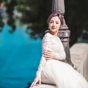 La mariée in a white lace wedding dress leans against a lamp post beside a body of water, with trees and blue water in the background. She has short, styled hair and a serene expression.