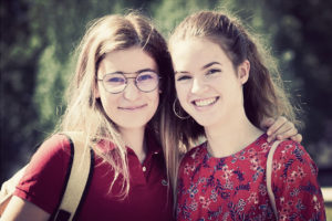 Two young women, embodying jeunesse, stand close together outdoors, smiling at the camera. The woman on the left has long hair, glasses, and is wearing a rouge polo shirt with a beige backpack. The woman on the right has her hair tied back, is dressed in a red floral top, and has hoop earrings.
