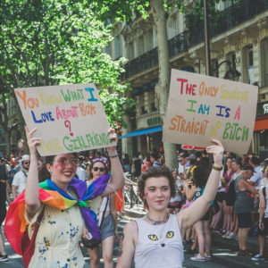 Two people at a pride parade hold colorful signs with LGBTQ+ messages. One sign says, "You know what I love about boys? Their girlfriend." The other says, "The only straight I am is a straight-up bitch.