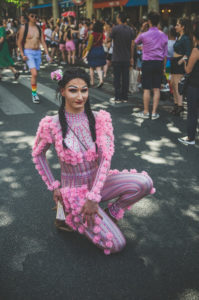 A person in a colorful, form-fitting pink outfit with flower details and braids poses on one knee at a lively outdoor event with a crowd in the background.