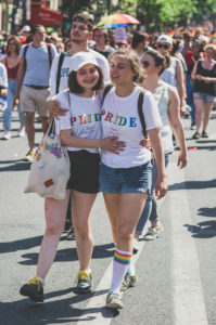 Two smiling young women wearing "PRIDE" shirts walk arm in arm at a pride parade, surrounded by a diverse crowd. One wears rainbow socks, the other a white cap. The scene is festive and colorful.