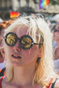 A person with light blonde hair and freckles wears large round sunglasses that read "L'AVEUGLE PAR AMOUR" at an outdoor event. A rainbow flag is visible in the background.