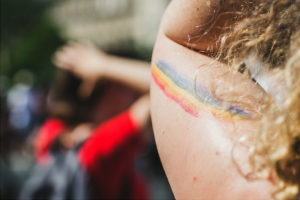 A close-up of a person's arm with a rainbow flag painted on it. The person has curly hair, and the background is blurred with people and outdoor sunlight visible.