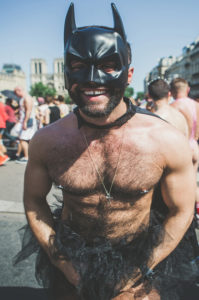A man wearing a black Batman mask, a black tutu, and a necklace smiles at an outdoor event. He is shirtless and appears to be enjoying a festive, sunny day with other people in the background.