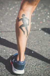 A close-up of a person’s lower leg showing a large tattoo of a running figure. The person is wearing a blue athletic shoe and standing on a textured pavement.