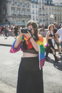 A person draped in a rainbow pride flag takes a photo with a camera at an outdoor event, with people walking and city buildings in the background.