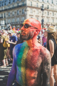 A bearded person with glasses and a bald head painted in rainbow colors stands shirtless in a crowd at an outdoor event, likely a Pride parade, with people and historic buildings in the background.