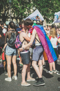 Two people embrace and kiss at a pride parade. One is wearing a rainbow flag as a cape. They are surrounded by other people celebrating, with flags and colorful outfits visible in the crowd.