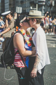 Two people embrace and share a kiss at an outdoor event. One wears a striped tank top and flower lei; the other wears a white shirt and hat. There are other people and buildings visible in the sunny background.