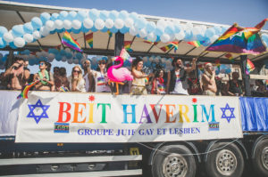 A colorful parade float decorated with balloons and rainbow flags carries people celebrating. A large banner reads, "Beit Haverim Groupe Juif Gay et Lesbien," with Stars of David and LGBT symbols visible.