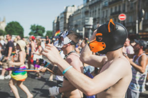 People in a parade wear colorful dog masks and minimal clothing, walking on a sunny city street. Rainbow flags and buildings are visible in the background, indicating a festive, inclusive event.