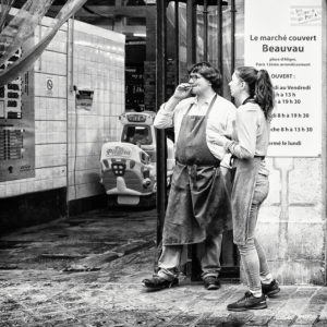 A black and white photo captures two people in aprons standing outside the marché Beauvau. One is drinking from a cup while the other is talking. A sign beside them reads "Le marché couvert Beauvau," with opening hours and location details. In the background, a children's ride adds a touch of whimsy to their pause.
