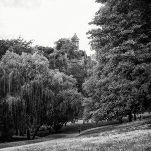 A black and white photo of a lush, tree-filled park with a small stone pavilion perched atop a rocky hill in the distance. A few people are seen strolling and relaxing on the grassy area in the foreground, capturing the serene atmosphere of Paris's Parc des Buttes-Chaumont.