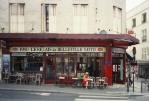 A person wearing a hat and red coat sits at an outdoor table of "PMU Le Relais de Belleville Loto," a charming corner café-restaurant. The urban setting features a red awning, several chairs, and tables. The light-colored building boasts shuttered windows.