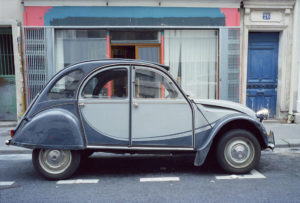 A vintage Citroën 2CV car is parked on a street in Ménilmontant, in front of a building with a blue door and a partially painted blue and pink facade. The car is a classic model with silver and blue tones.