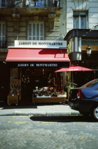 A sidewalk view of the small shop, Jardin De Montmartre, in Paris. The Paris attraction features a red awning and a display of fresh fruits and vegetables outside. An adjacent red patio umbrella shades the storefront, while a black car is parked on the cobblestone street.