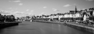 A black-and-white panoramic view of the waterfront town of La Touques, with a river running through it. Lined with buildings, trees, and people, the townscape includes a prominent church steeple and a cloudy sky.