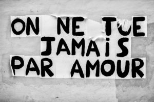 A black and white image features French text pasted on a wall. The text reads "ON NE TUE JAMAIS PAR AMOUR," which translates to "One never kills for love" in English. The bold, slightly irregular letters enhance the handmade appearance, emphasizing the poignant message of amour and its innocence juxtaposed against violence.